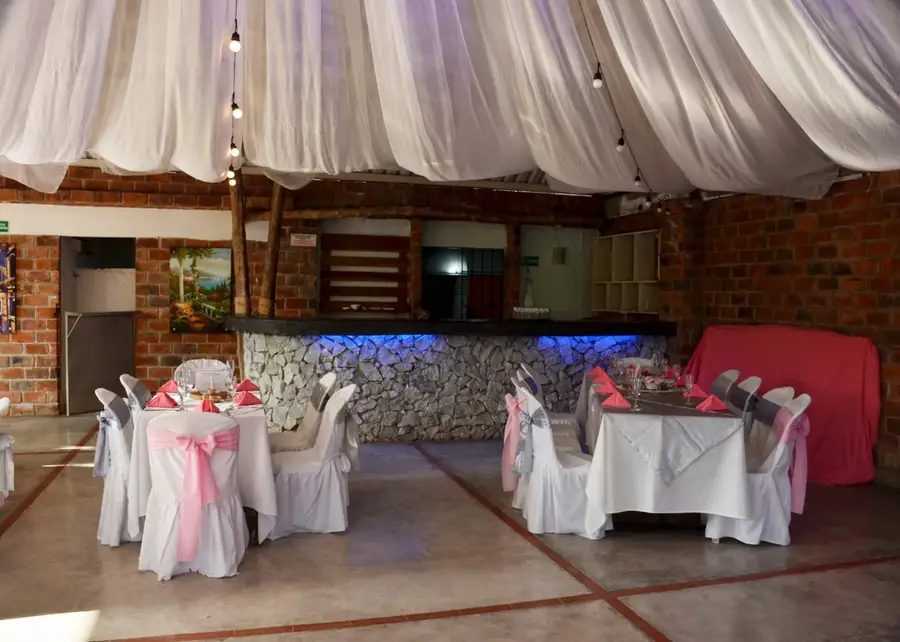 Salón montado para un evento: mesas vestidas de blanco con lazos rosados, techo de telas con bombillos y la barra de piedra al fondo